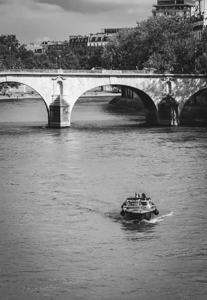 Black and white view of a small boat cruising on the Seine River beneath a stone bridge in Paris