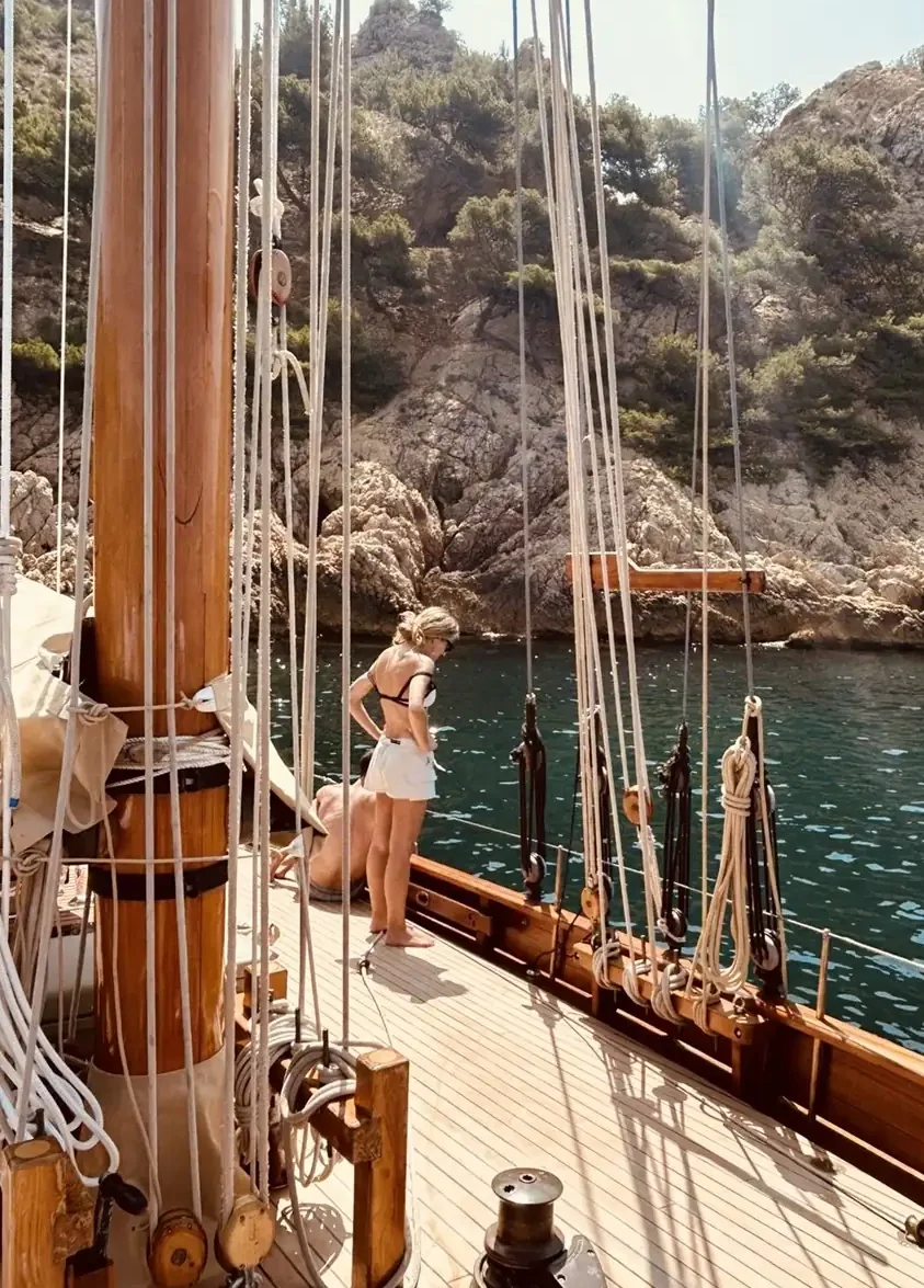 Woman standing on the deck of a sailboat looking out at the lush Mediterranean coastline