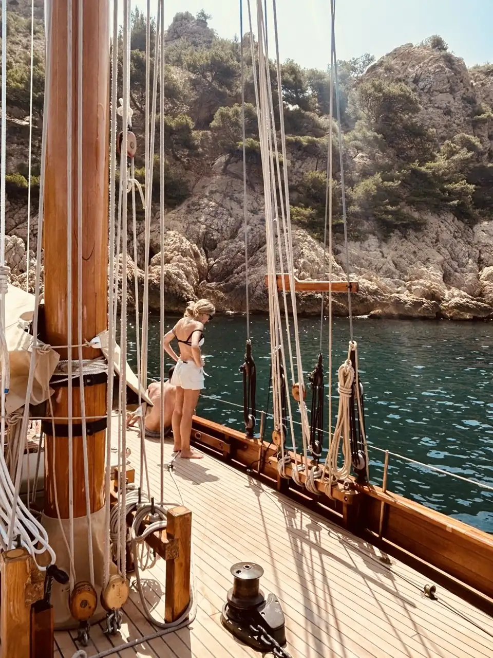 Woman standing on the deck of a sailboat looking out at the lush Mediterranean coastline