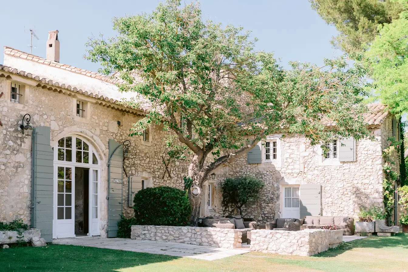 Exterior facade of a traditional stone farmhouse in Provence shaded by a large tree