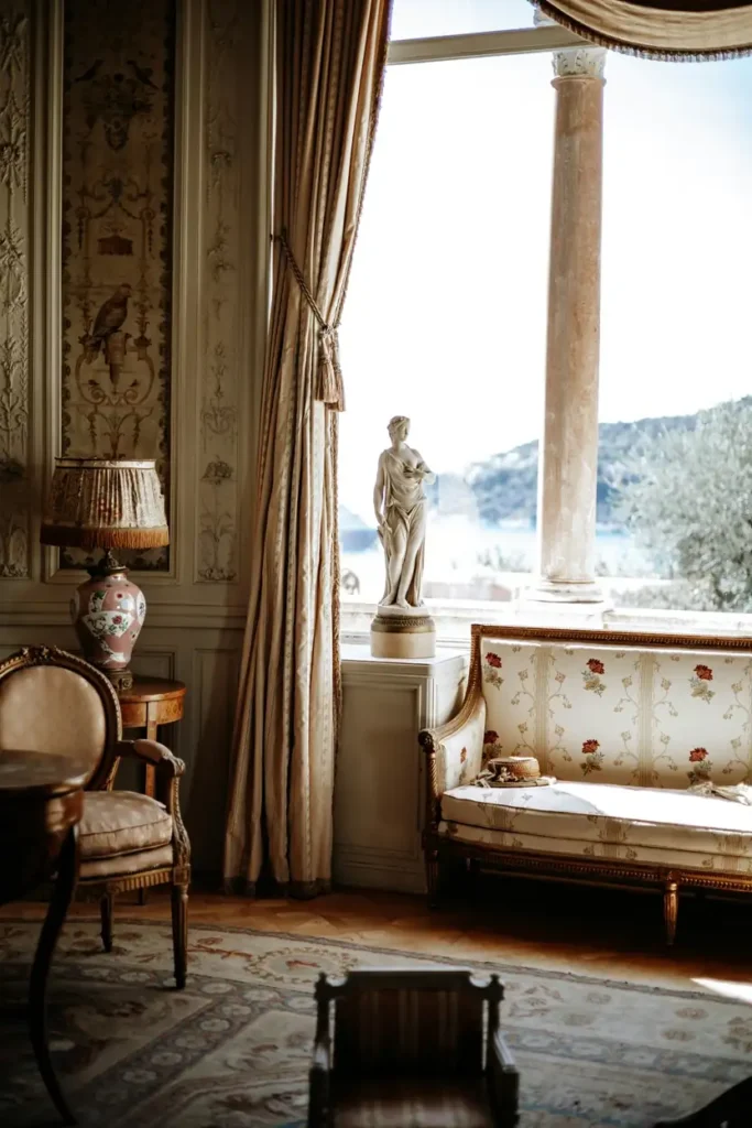 Elegant vintage bedroom with draped curtains, antique furniture, and large window overlooking scenic view