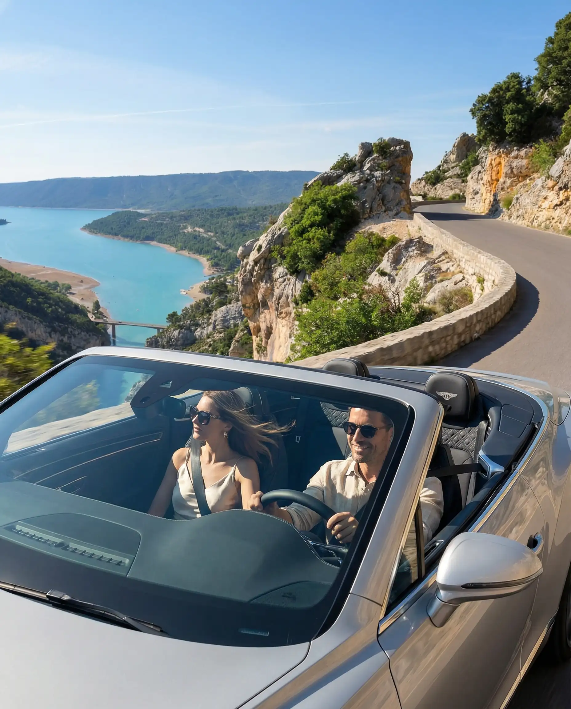 Chic couple driving a silver luxury convertible on a winding coastal road overlooking the Verdon Gorge turquoise water.