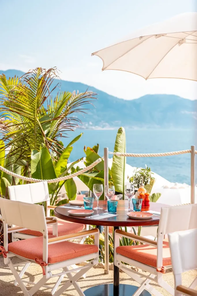 Alfresco dining area at Maybourne La Plage with coral-cushioned chairs, white umbrellas, and tropical greenery overlooking the coast.