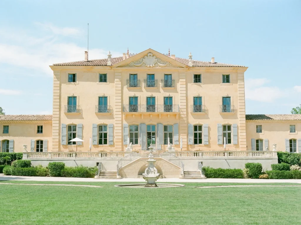 Front facade of a grand neoclassical yellow chateau in France with blue shutters and a formal garden.
