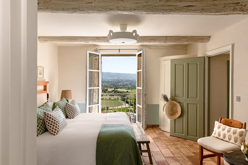 Elegant rustic bedroom in a French chateau with open balcony doors overlooking a lush vineyard.