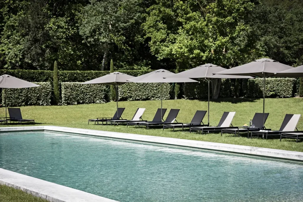 Large outdoor swimming pool surrounded by chaise lounges, sun umbrellas, and a lush green forest