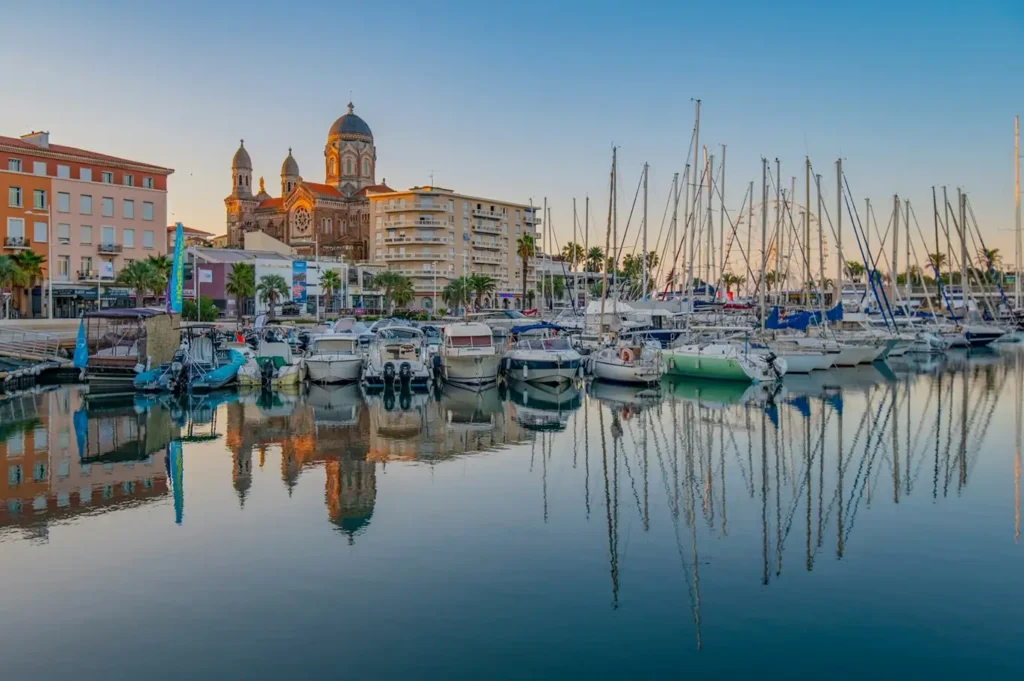 Luxury yachts moored in Saint-Raphaël marina with the Basilique Notre-Dame de la Victoire in the background at sunset.