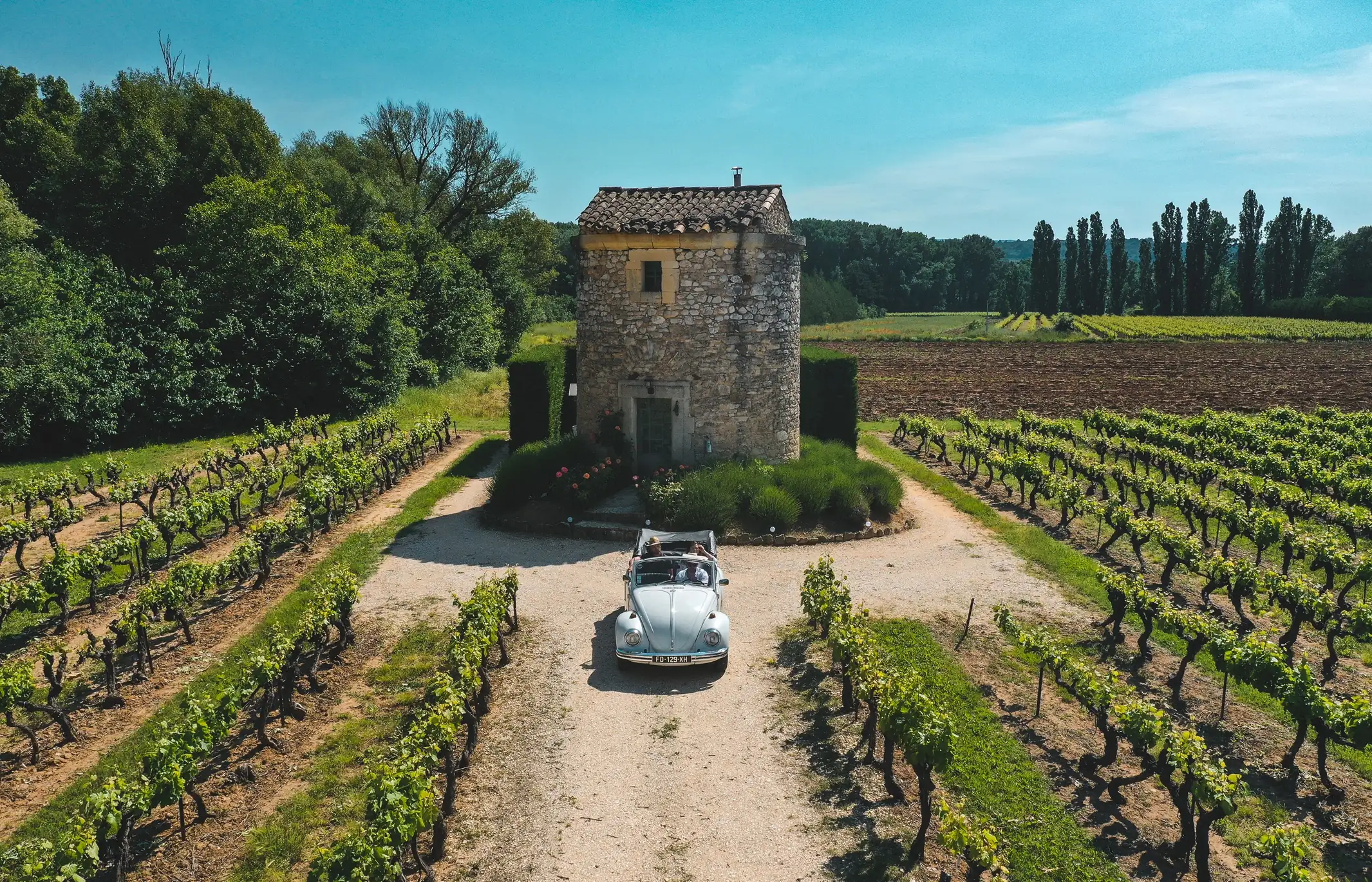 Vintage white Volkswagen Beetle convertible parked in front of a stone pigeonier cottage in a lush Provence vineyard.