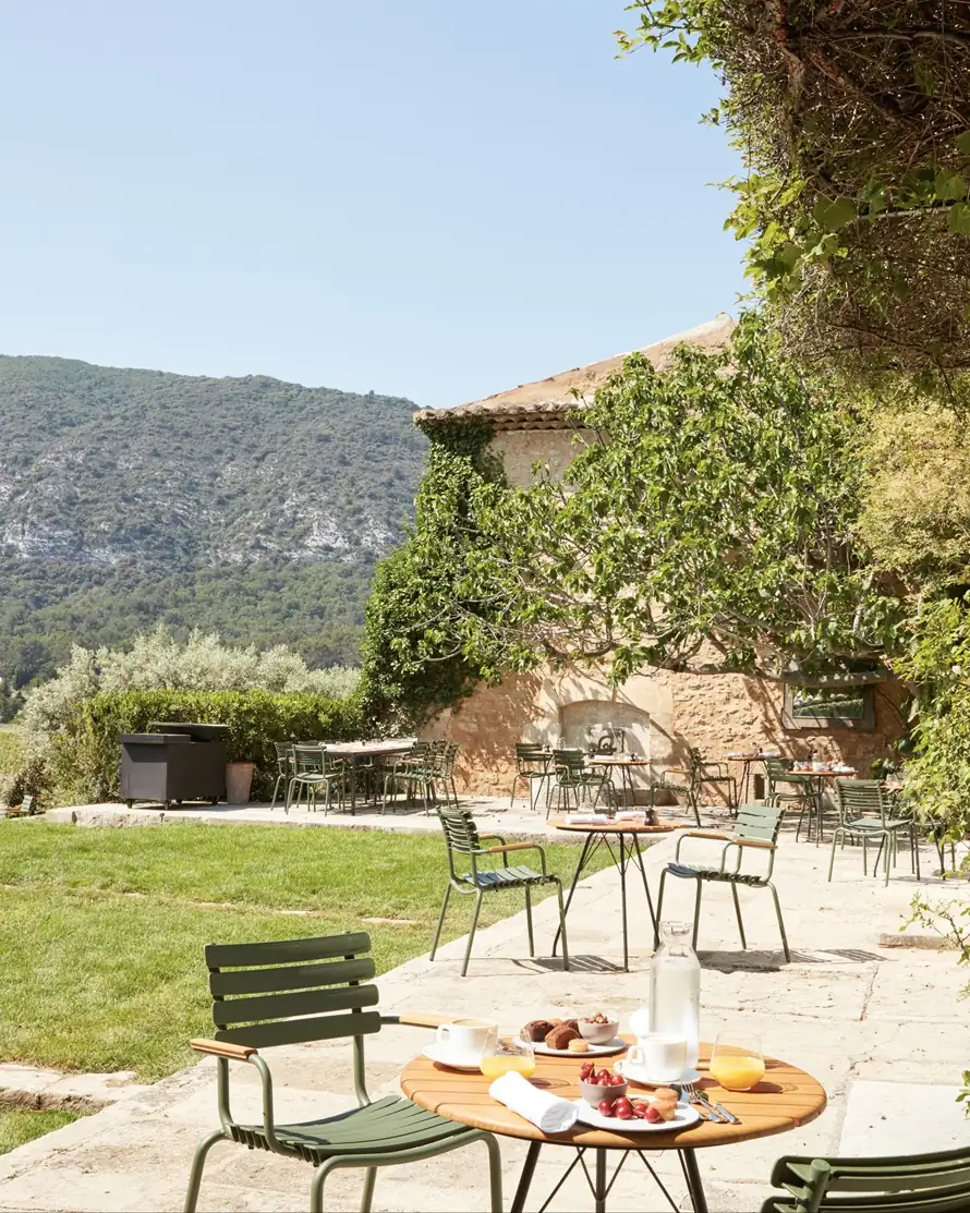 Green bistro chairs and tables on a sunlit stone terrace before a Provence bastide and olive trees